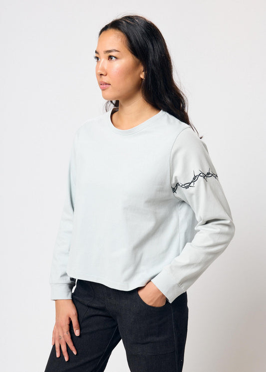 Woman wears cropped pale blue long sleeve t-shirt with black embroidered barbed wire design on left arm