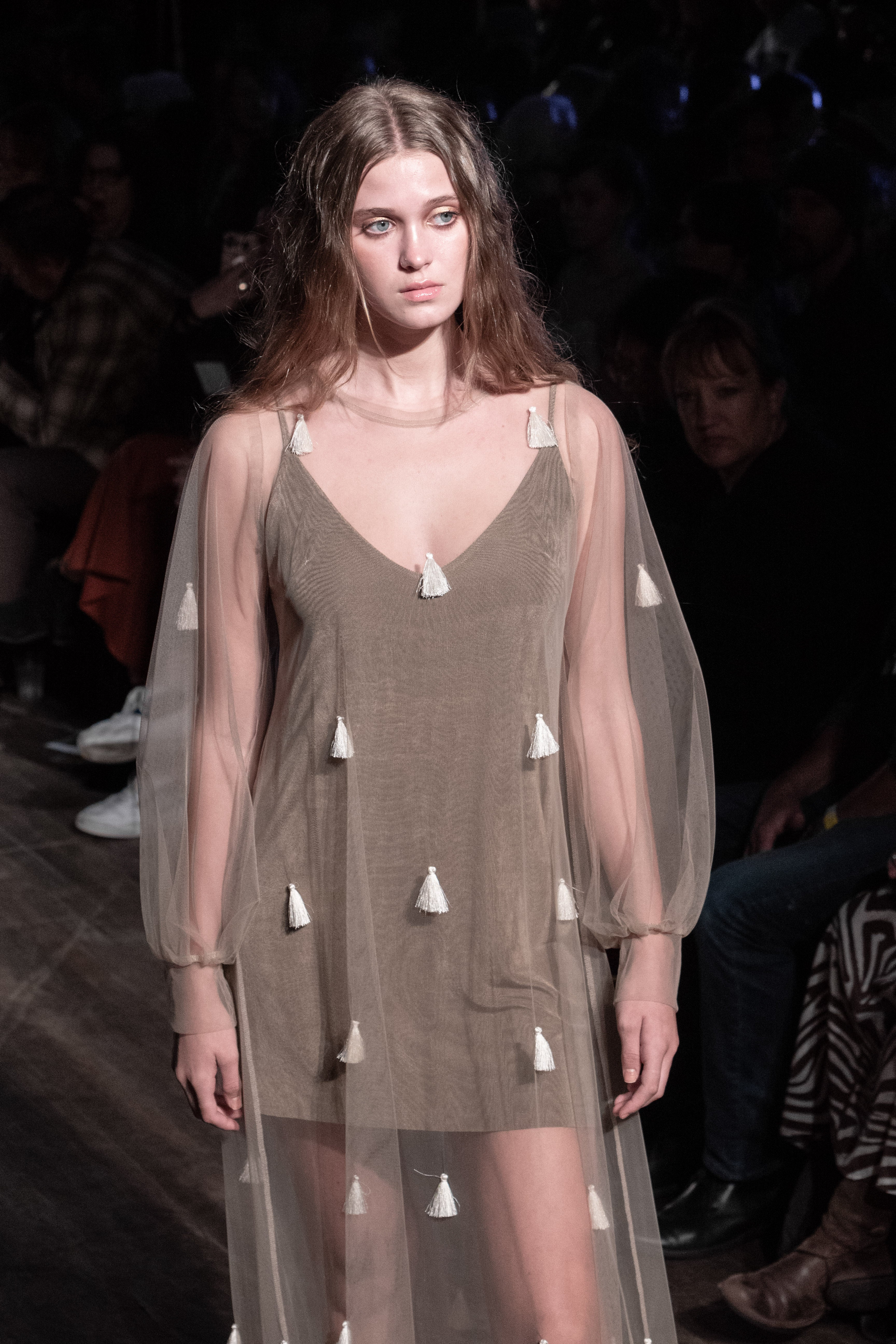 Model wearing a green dress with sheer overlay and white tassels on a runway.
