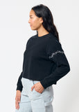 Woman wearing cropped black long sleeve tee shirt with embroidered gray barbed wire design on left arm