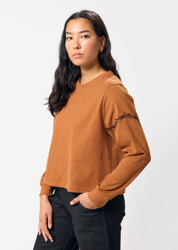 Woman wearing a brown long-sleeve top with embroidered black barbed wire detail on sleeve on a white background