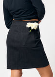 Person wearing a black skirt with a white flower in the pocket on a plain background