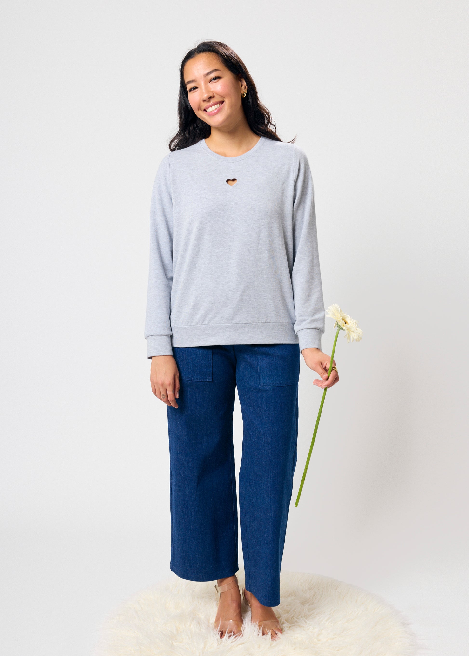 Woman wearing a light gray sweatshirt with a heart cut out and blue pants holding a flower on a white background