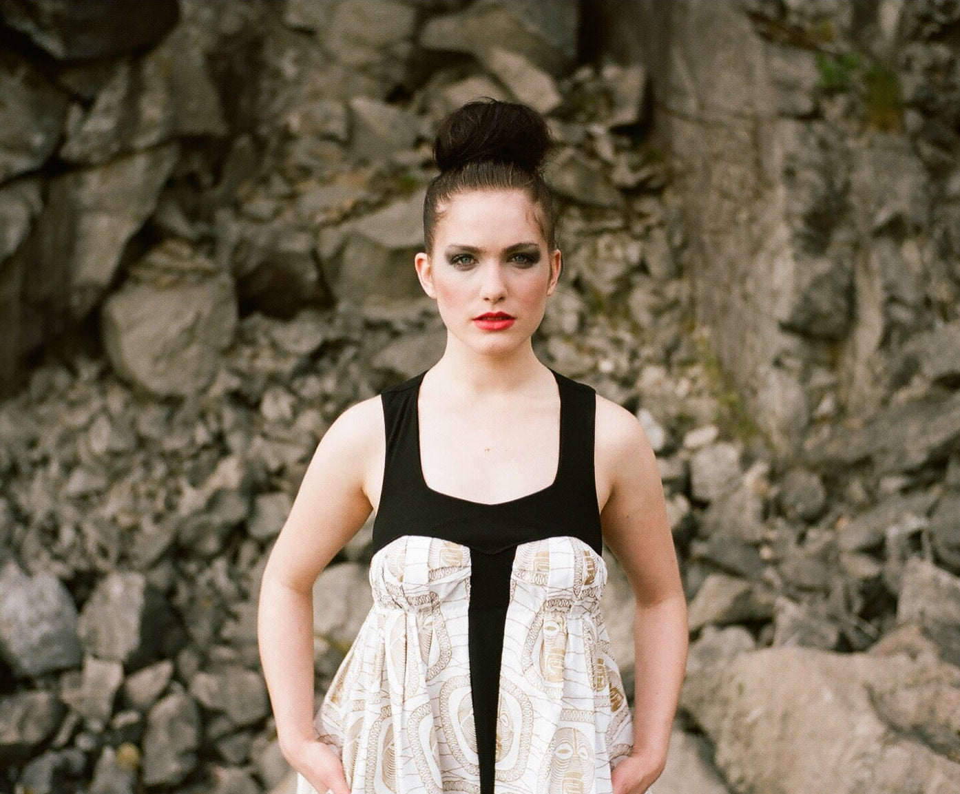 Woman in a black top and patterned skirt standing against a stone wall.