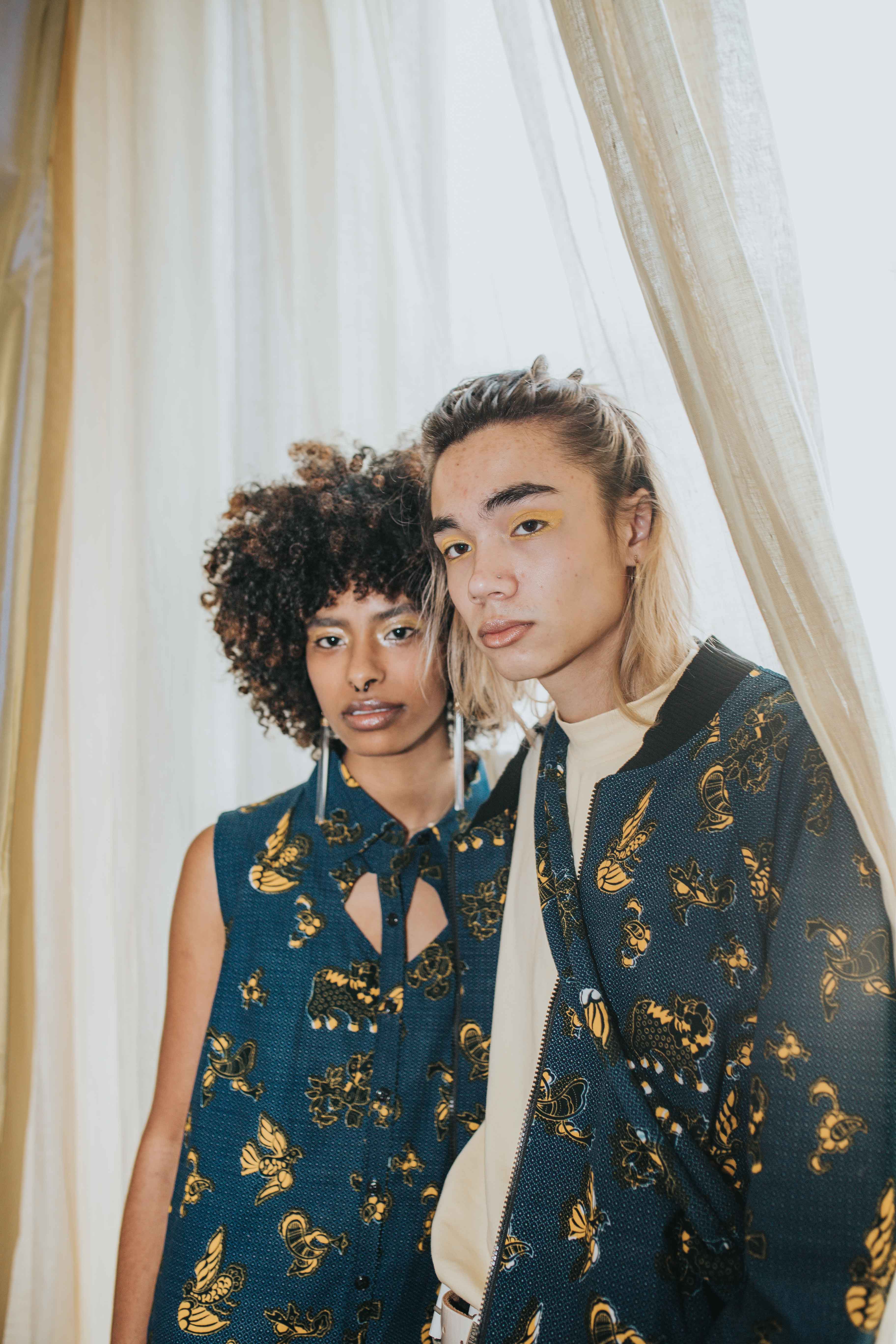 Two individuals wearing matching dark blue outfits with gold floral patterns in front of a white curtain.