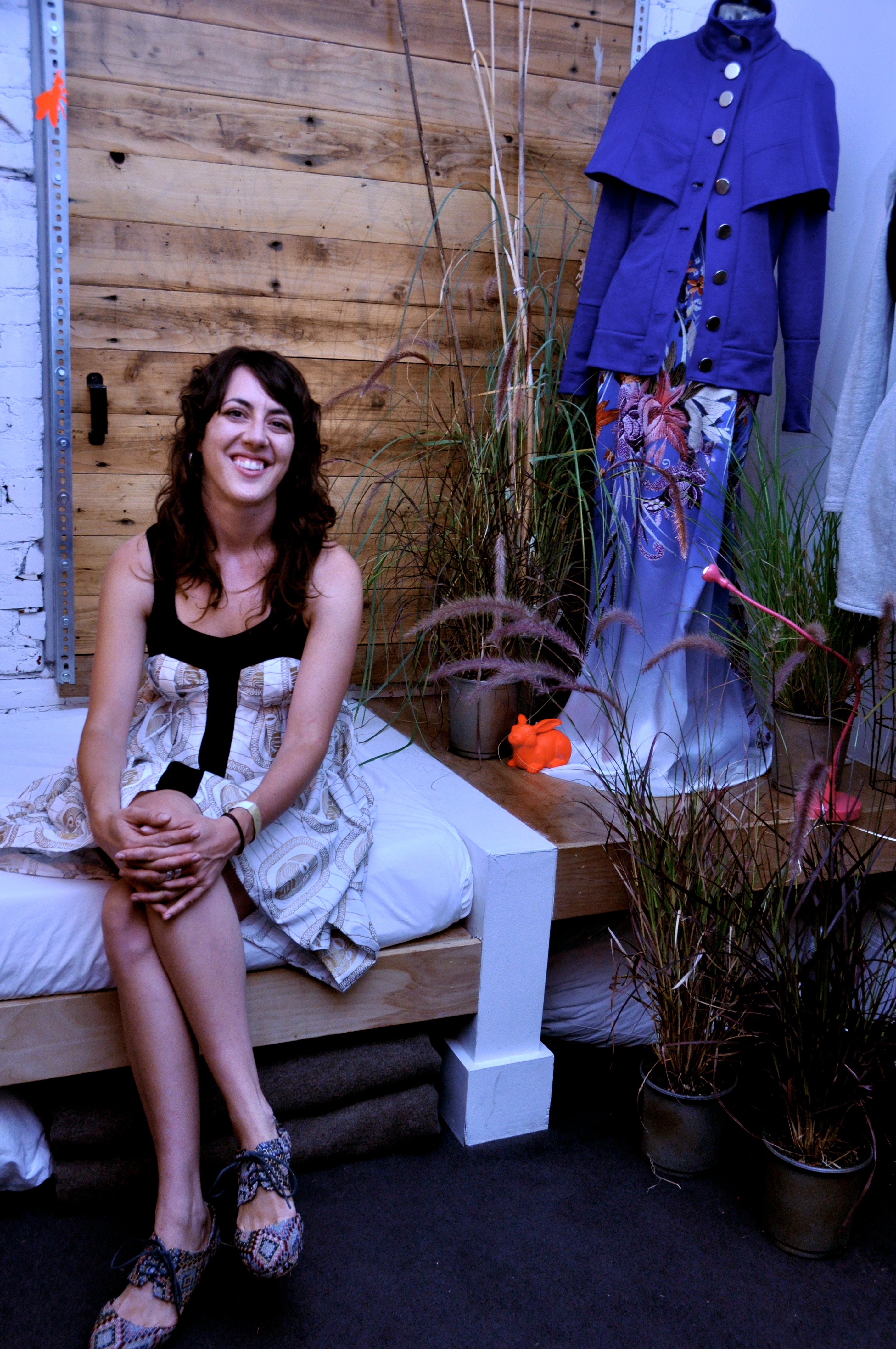 Woman sitting on a wooden bench with plants and a purple coat in the background