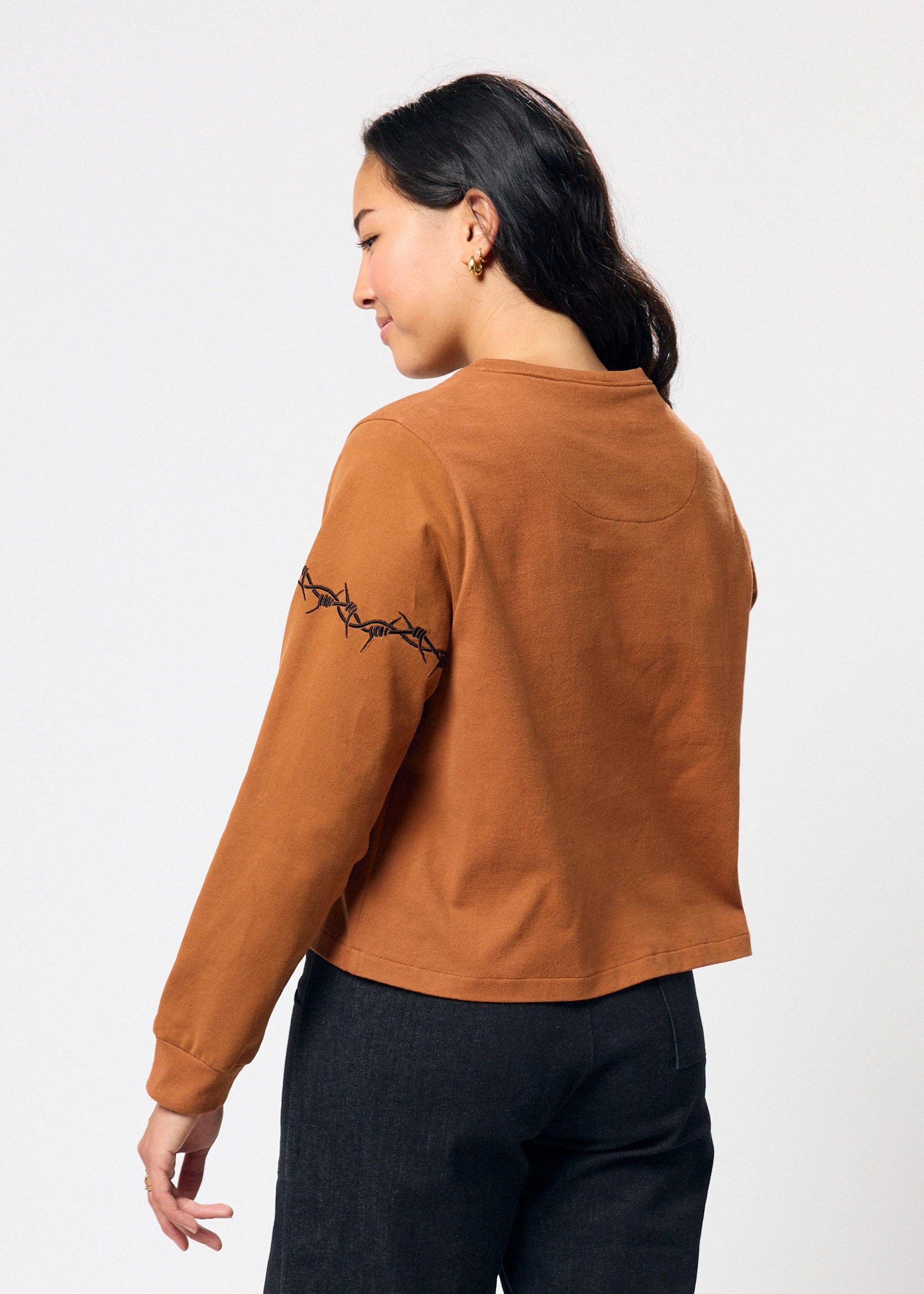 Woman wearing a brown long-sleeve top with embroidered black barbed wire detail on sleeve on a white background
