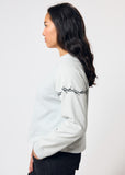Woman wears cropped pale blue long sleeve t-shirt with black embroidered barbed wire design on left arm
