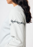 Woman wears cropped pale blue long sleeve t-shirt with black embroidered barbed wire design on left arm