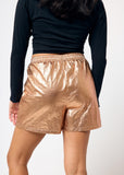 Person wearing a black long-sleeve top and bronze metallic shorts on a white background