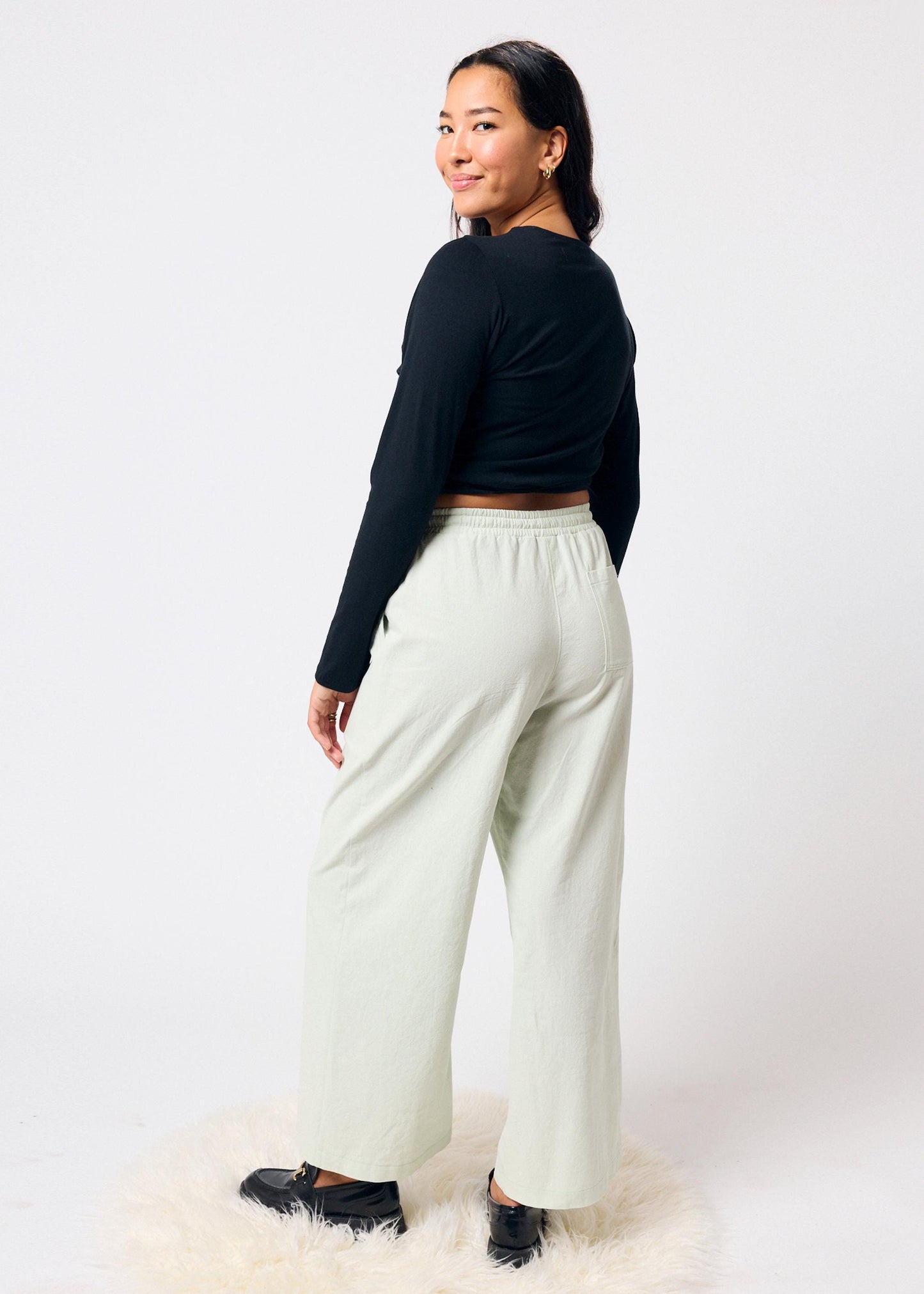 Woman wearing a black long-sleeve top and pale green colored pants on a white background