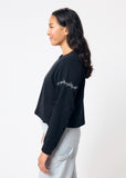 Woman wearing cropped black long sleeve tee shirt with embroidered gray barbed wire design on left arm