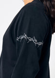 Woman wearing cropped black long sleeve tee shirt with embroidered gray barbed wire design on left arm