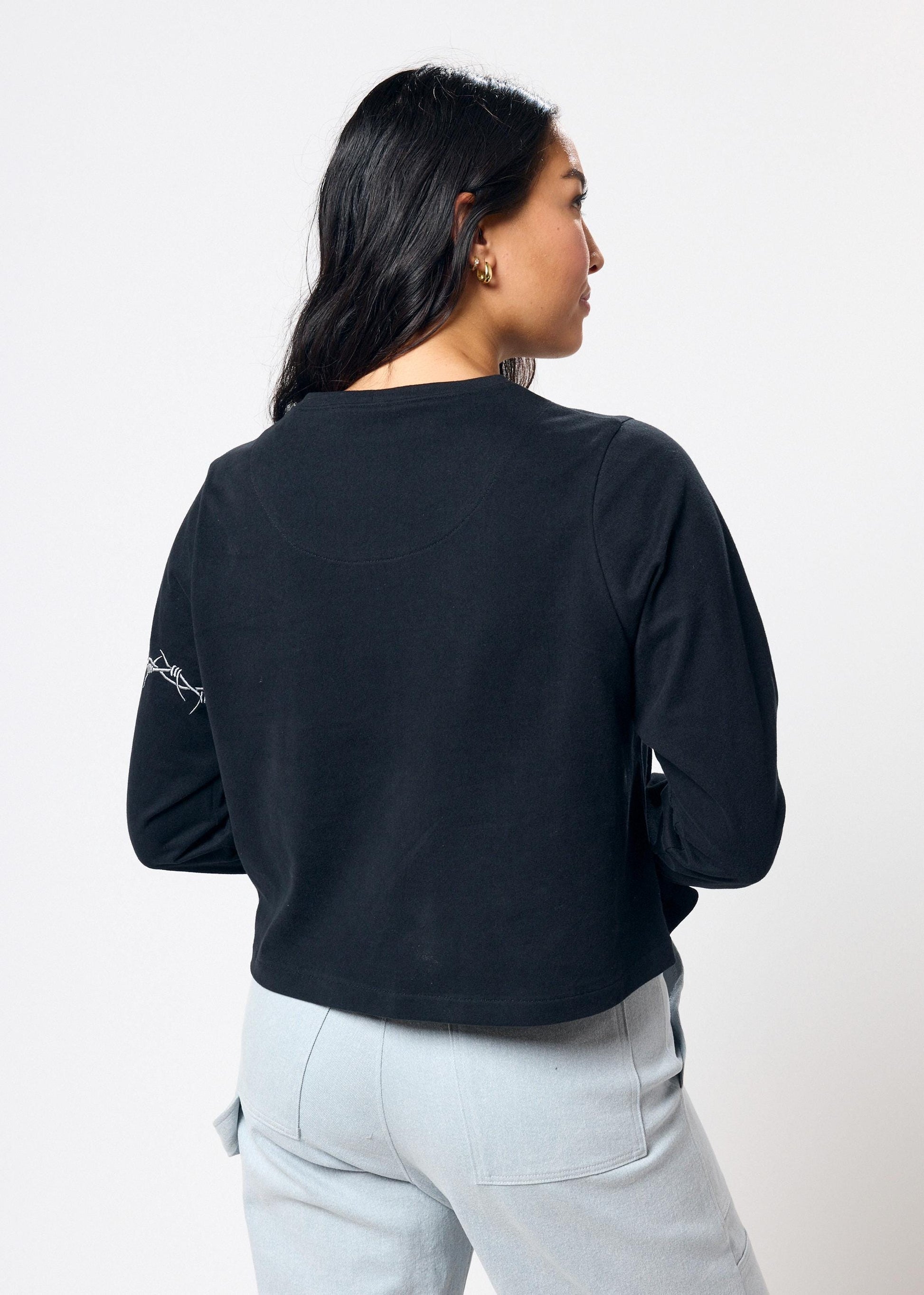 Woman wearing cropped black long sleeve tee shirt with embroidered gray barbed wire design on left arm