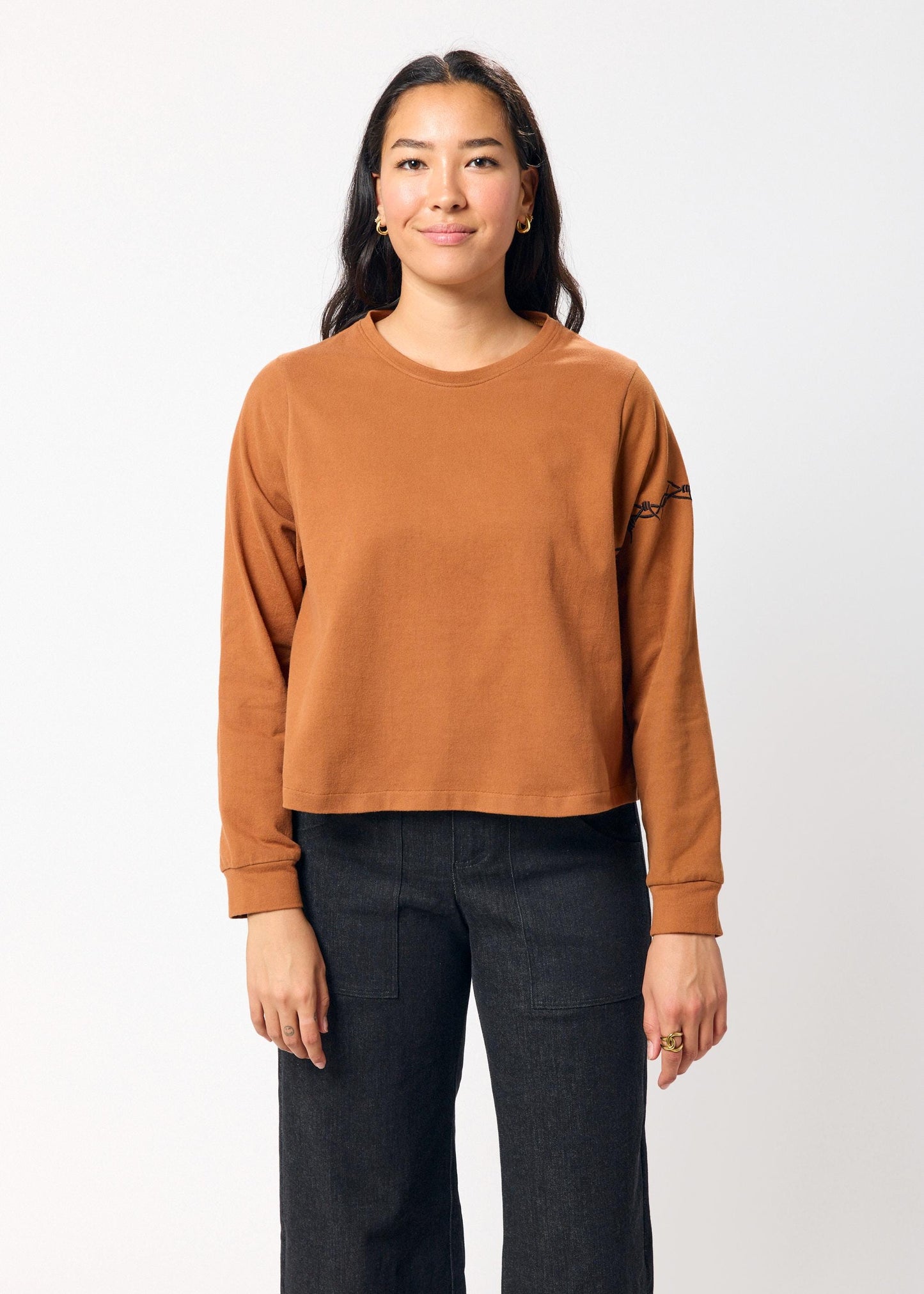 Woman wearing a brown long-sleeve top with embroidered black barbed wire detail on sleeve on a white background