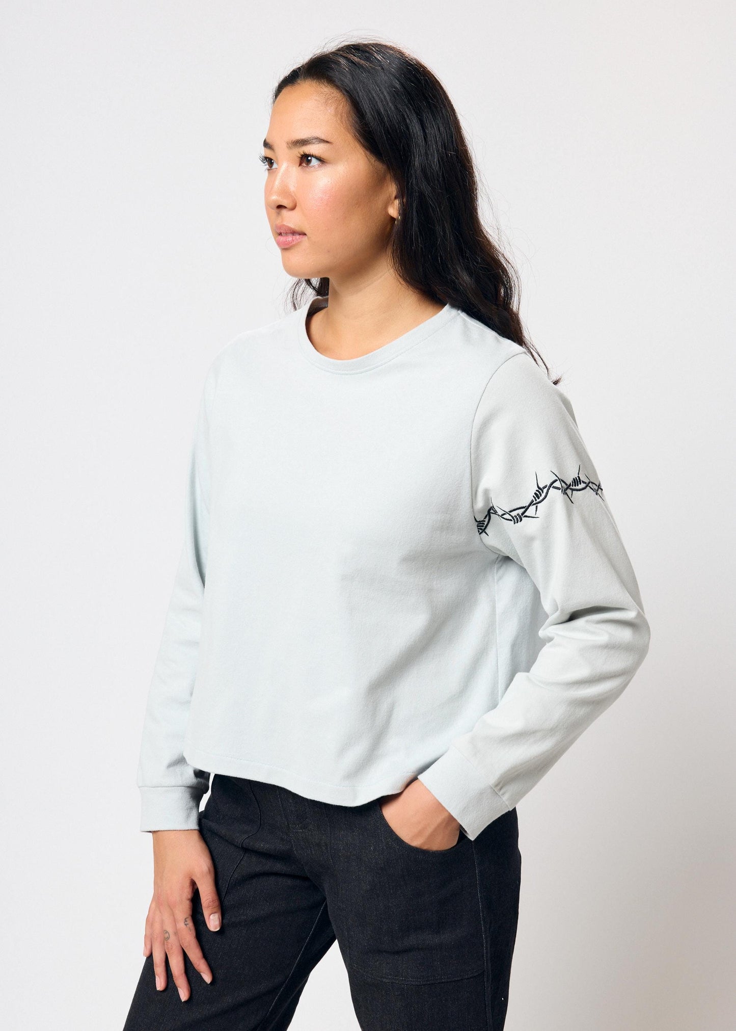 Woman wears cropped pale blue long sleeve t-shirt with black embroidered barbed wire design on left arm
