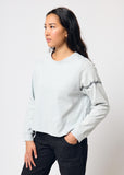 Woman wears cropped pale blue long sleeve t-shirt with black embroidered barbed wire design on left arm